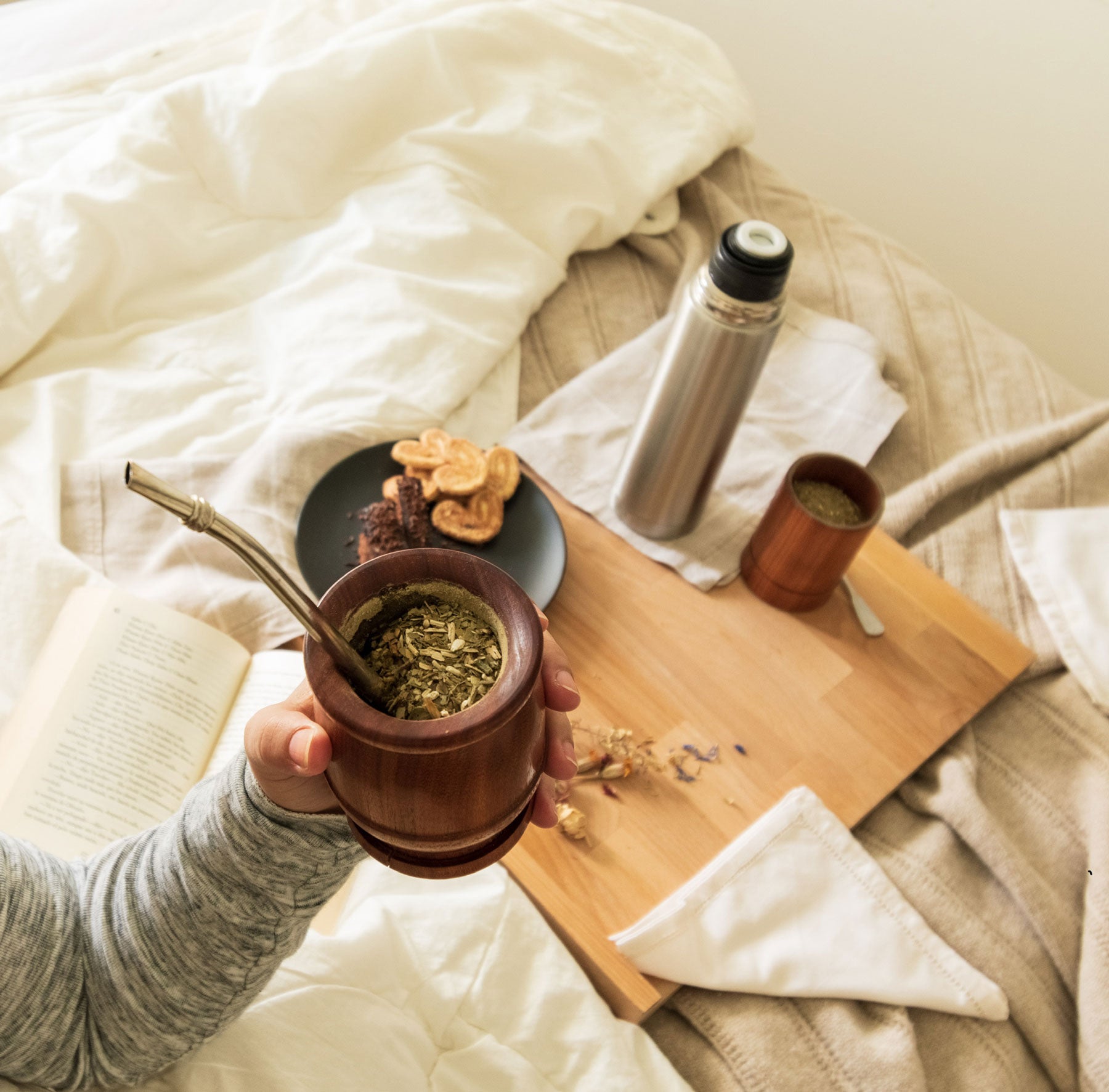 Person holding a wooden mate cup with a hot beverage on a bed with a wooden tray and snacks.