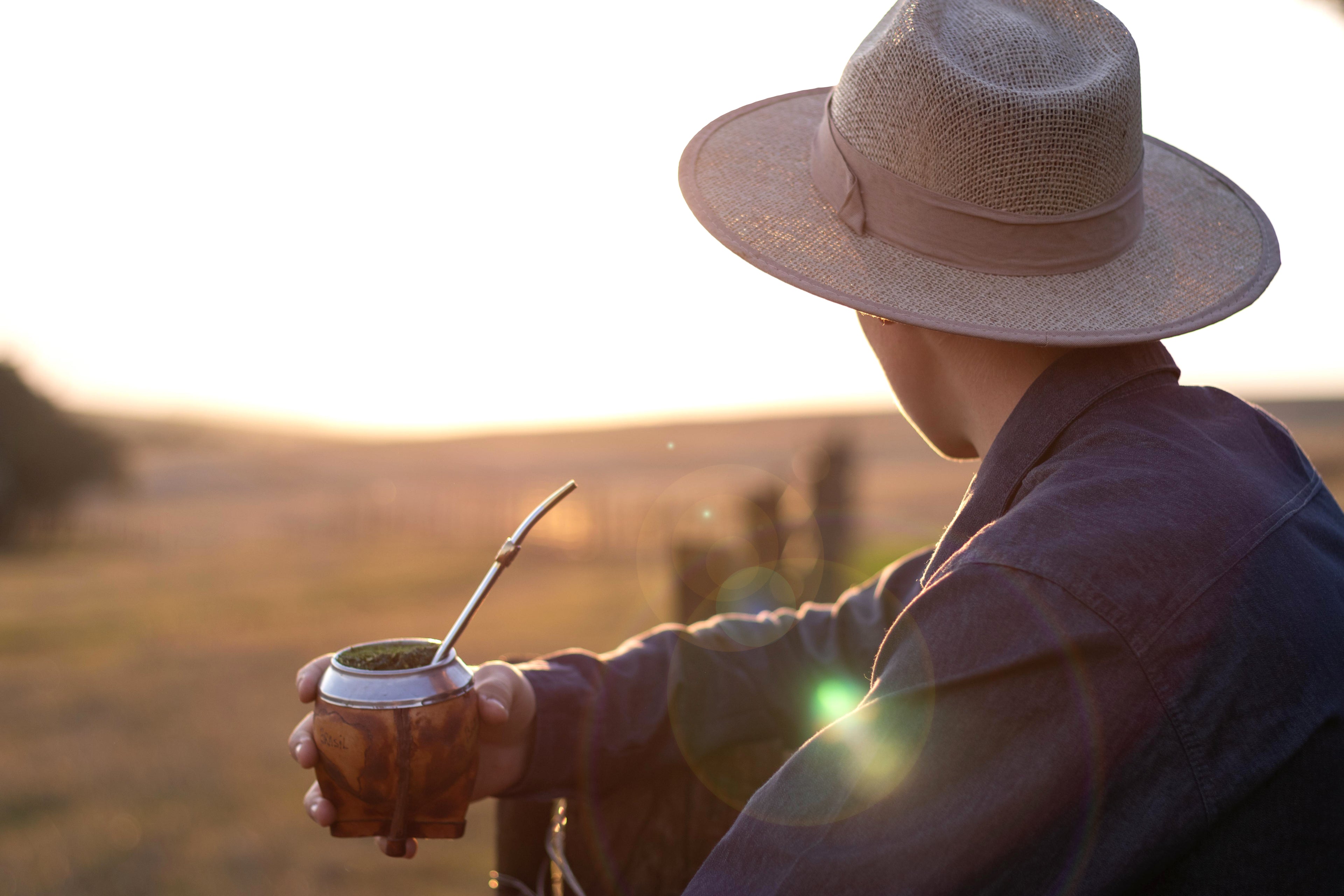 Person in a straw hat holding a mate gourd with a bombilla in a field at sunset